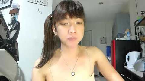 Video of an Asian woman with long brown hair in a ponytail, wearing a spaghetti-strap tank top and a pendant necklace. She's in a cluttered, dimly-lit room with a motorcycle, a fridge, and various personal items in the background.