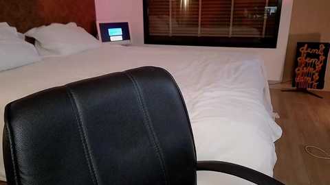 Media: Video of a modern bedroom with a white bed, black leather chair, wooden floor, and large window with blinds. A digital clock and a framed sign are visible.