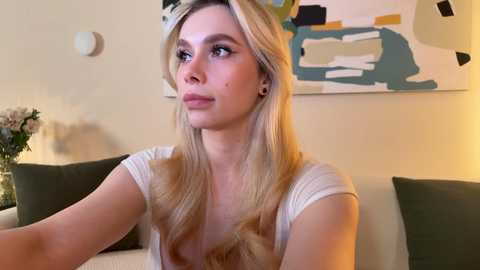 Media: A video of a blonde Caucasian woman with long hair, wearing a white top, seated on a beige couch. Behind her, an abstract painting and a vase with flowers.