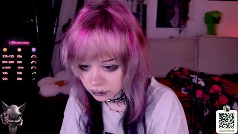 A young woman with pastel pink and purple hair and heavy makeup in a dimly lit room, wearing a black choker, white shirt, and a black and red plaid blanket.