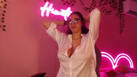 Media: Video of a young woman with light skin and dark hair, wearing glasses and a white button-up shirt, posing with arms raised in a pink-lit room with star and heart neon lights.