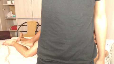 Video of a nude person lying on a bed in a bedroom, wearing only a black tank top, with a wooden chair and a closet in the background.