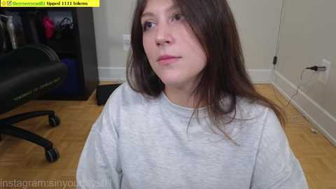 A video of a young Caucasian woman with long brown hair, wearing a gray sweatshirt, sitting on a wooden floor. Background includes a black gaming chair and a partially visible white wall.