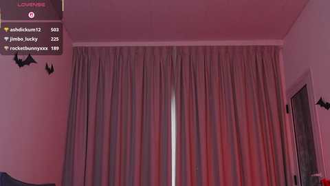Video of a dimly lit bedroom with a tall, closed window covered by beige curtains. The room has a reddish hue, and a small, black bat decoration hangs on the left wall.