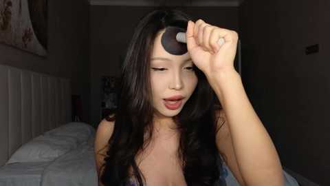 A video of a young Asian woman with long black hair, wearing minimal makeup, using a black eyeshadow brush to touch her eye in a dimly lit bedroom.