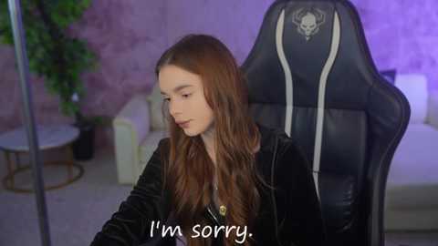 Video of a young girl with long, wavy brown hair, wearing a black velvet jacket, sitting in a black gaming chair with a skull emblem, in a softly lit room with purple walls. Text overlay reads, \"I'm sorry.\