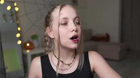 Media: Video of a young woman with fair skin, platinum blonde hair, and a black tank top. She has a relaxed, open-mouthed expression in a dimly lit room with string lights and a beige couch in the background.