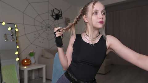 Video of a young woman with light skin, blonde braided hair, wearing a black tank top and black fingerless gloves, holding a hairbrush, in a modern, dimly-lit room with a spiderweb wall decoration and a mirror with warm lights.