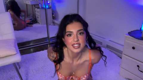 Video of a young woman with long dark hair, fair skin, and full lips, wearing a pink and orange floral bra, kneeling on a plush white carpet in a softly lit bedroom with mirrors and white furniture.