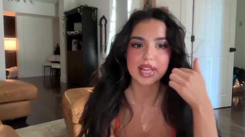 Video of a young woman with long, wavy black hair, light brown skin, and full lips, wearing a revealing red dress, in a modern living room with beige furniture, a dark cabinet, and a door.