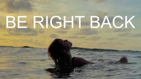 Video of a silhouette of a woman with wet hair, lying on her back in shallow ocean water, facing the sunset. The sky is a mix of golden and blue hues, with the words \"BE RIGHT BACK\" in bold white text overlaid.