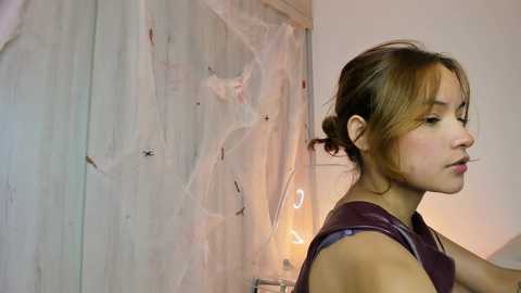 Video of a young woman with light skin and auburn hair styled in a messy bun, wearing a sleeveless purple top. She's indoors with a blurred, abstract painting in the background.