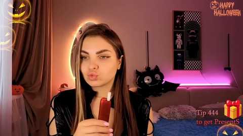 Video of a young woman with long brown hair, wearing a black outfit, pouting with a lollipop in her mouth, in a Halloween-themed bedroom with decorations and a black cat.