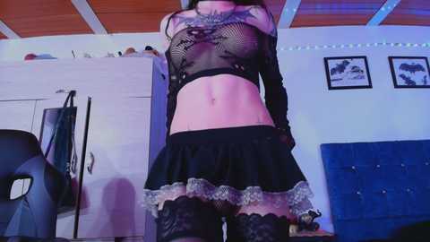 Video of a slender woman with pale skin, wearing a black fishnet top and short skirt, standing in a room with white walls, wooden ceiling, and framed art.