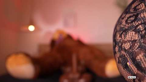 Media: Video of a blurry, intimate scene featuring a woman with dark skin and curly hair, wearing black lace lingerie, lying on a bed with warm lighting and soft pink decor in the background.