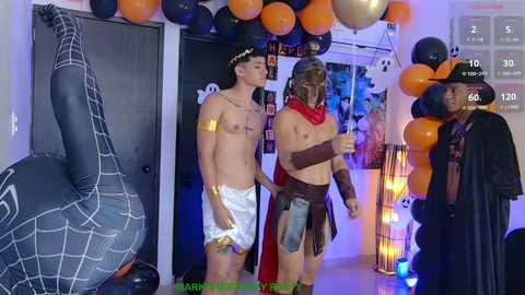 Media: Video of a Halloween party with two shirtless young men in white shorts and costumes, one in a red cape, and a man in a black cape. Spiderman and Spiderman balloons hang from the ceiling, with a wall of spider webs in the background.
