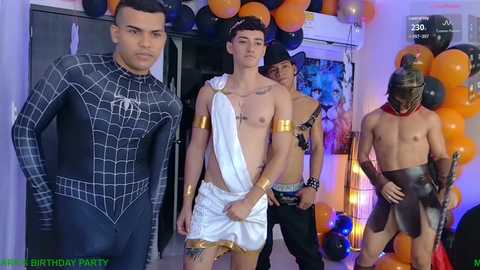 Video of three young men in superhero and Greek god costumes at a birthday party. One in Spider-Man suit, another in white toga, and a third in black leather. Background features orange balloons, a poster, and a cake.