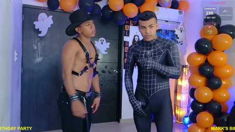 Media: Video of two shirtless, muscular men in black harnesses and hats, standing in a Halloween-themed room with orange balloons and a Spider-Man costume, in a party setting.