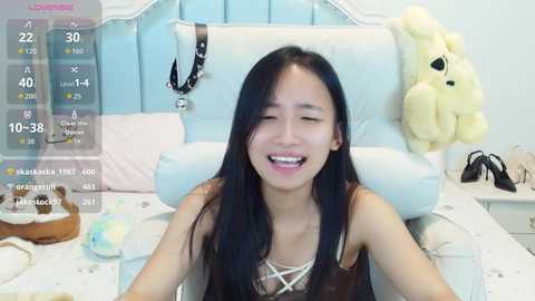 Video of an Asian woman with long black hair, wearing a black lace top, smiling joyfully in a pastel-colored bedroom with a white upholstered headboard and plush toys.