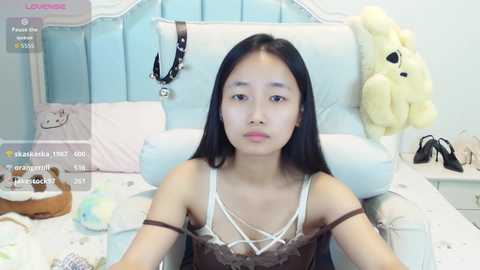 Video of an Asian woman with long black hair, wearing a brown lace camisole, sitting on a white bed with a stuffed bunny, in a light-colored room.