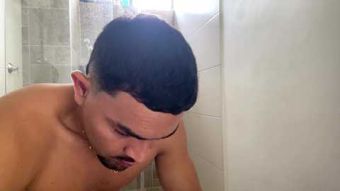 Video of a shirtless man with short dark hair, in a tiled shower, bending over and looking down, water streaming from his hair.