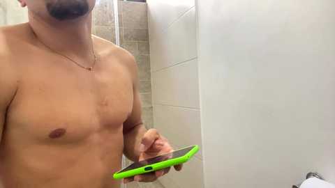 Video of a shirtless, light-skinned man with a trimmed beard, wearing a thin gold chain necklace, holding a bright green smartphone in a white-tiled bathroom.