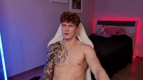 Video of a shirtless, fair-skinned young man with curly red hair, one sleeve covered in a detailed tattoo, sitting on a gaming chair in a dimly lit bedroom with red lighting, black bedding, and a framed picture on the wall.
