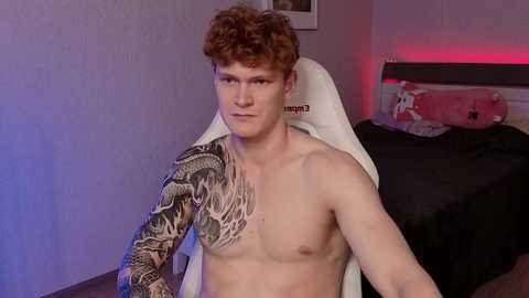 Video of a young, shirtless man with curly red hair and a large, intricate black and gray tattoo on his left arm, seated on a white chair in a dimly lit bedroom with a pink pillow and a bed with black sheets.