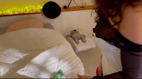 A video of a woman with curly brown hair wearing a black top, standing next to a bed with a white quilt, a gray stuffed bear, and a window with a yellow light behind it.