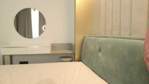 Video of a minimalist bedroom with a circular mirror, light gray upholstered headboard, white bed, and cream curtains.