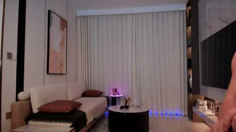 Video of a modern, minimalist living room with beige walls, white curtains, a white sofa, and a round black coffee table. Artwork, decorative items, and ambient lighting add a serene ambiance.