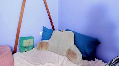 Video of a cozy corner in a child's room featuring a green Minecraft Steve plush, a plush kangaroo, and blue pillows on a light bedspread against a lavender wall with wooden triangle shelves.