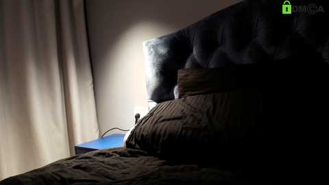Video of a dimly lit bedroom featuring a dark, tufted headboard, a beige blanket, and a small lamp emitting a warm glow on the left side.