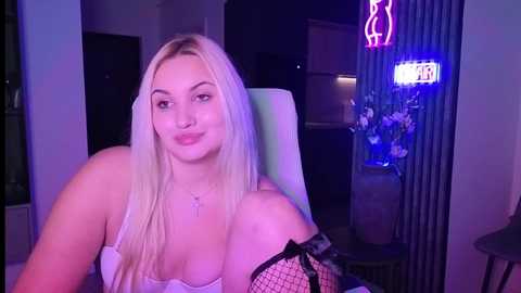 A video of a young, fair-skinned, blonde woman with long hair, wearing a white camisole and black fishnet sleeves, sitting in a dimly lit room with neon signs and a vase of flowers in the background.