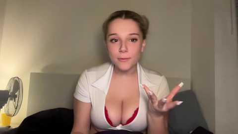 Video of a young Caucasian woman with light skin, brown hair, and large breasts, wearing a white cropped shirt revealing a red bra, sitting on a bed with a beige headboard.