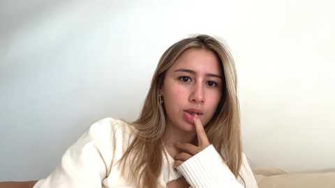Video of a young woman with long, straight, blonde hair, light skin, wearing a white sweater, holding her index finger to her lips, against a plain, light-colored background.