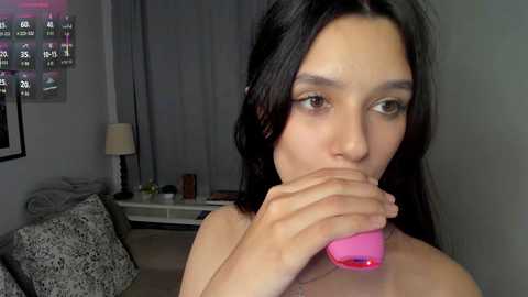 A video shows a young woman with long black hair, wearing a pink vibrator, in a dimly lit living room with gray curtains, a patterned sofa, and a lamp.