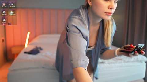 Media: Video of a blonde woman in a light blue robe playing a video game on a bed with a pink lamp in the background.
