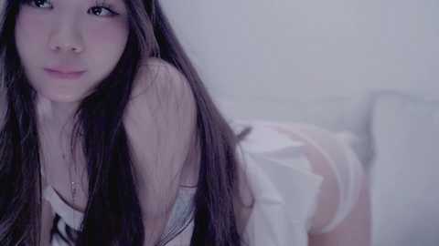 A soft-focus video of an Asian woman with long black hair, light skin, and a delicate facial structure, wearing a white lace bra and panties, lying on a white bed with a blurred background.