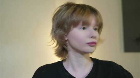 Video of a young Caucasian boy with light brown, shoulder-length hair, wearing a black shirt, standing against a beige wall with a dark rectangular object in the background.