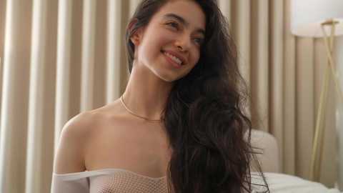 Video of a young woman with long, wavy dark hair, fair skin, and a slender physique, wearing a white, off-shoulder dress, standing against beige curtains in a softly lit room.