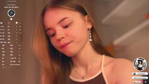 Media: Video of a young Caucasian woman with long brown hair, wearing a sleeveless top and dangling earrings, smiling softly. Background shows a blurred, indoor setting.