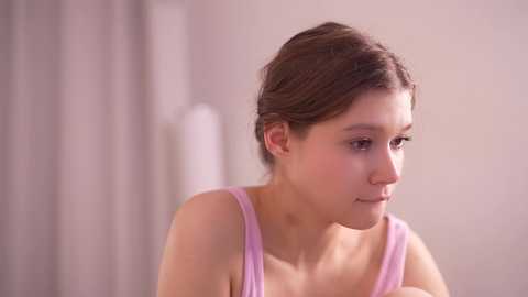 Video of a young Caucasian woman with light skin and brown hair, wearing a light pink tank top, looking contemplative against a softly lit, out-of-focus background.