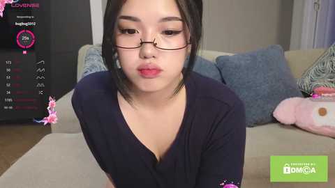 A video of an East Asian woman with a fair complexion, wearing glasses and a navy-blue top, sitting on a beige couch. Background shows a TV screen displaying live chat statistics, and a plush toy.
