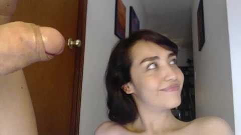 Video of a young woman with dark hair, pale skin, and a small bust, smiling at a large, erect penis in a doorway.