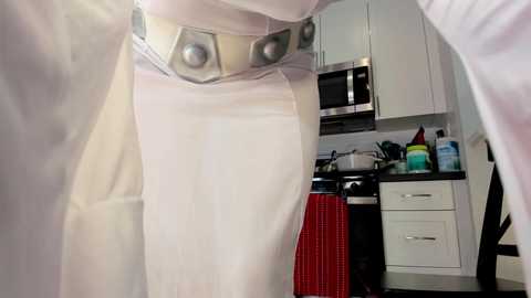 A video of a woman in a white lab coat with silver buttons, standing in a modern kitchen with white cabinets, stainless steel appliances, and red and black accents.