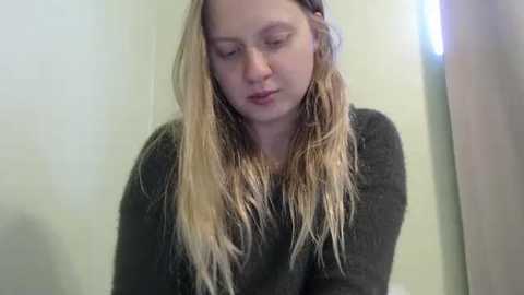 Media: Video of a young, fair-skinned woman with long, wet, blonde hair, wearing a dark gray sweater, looking down pensively against a pale green wall.