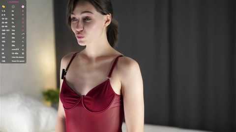 Video of a young woman with fair skin and dark hair, wearing a sheer red lingerie top, standing in a dimly lit room with dark curtains in the background.