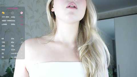 A video of a blonde woman with fair skin, wearing a white off-shoulder dress, her head slightly tilted back, lips parted, in a brightly lit room with floral wallpaper.