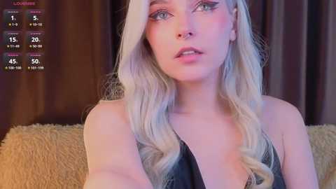 Video of a young, fair-skinned woman with long, wavy blonde hair, wearing a black top, looking pensively towards the camera. Background features a beige couch and brown curtains.